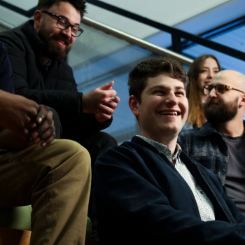 Group of young professionals sitting on indoor stairs, smiling and engaging in conversation in a modern office setting.